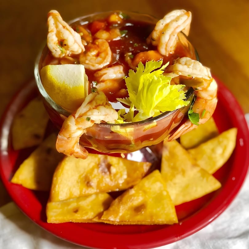 Shrimp cocktail in a glass with lemon and celery, served with tortilla chips on a red plate.