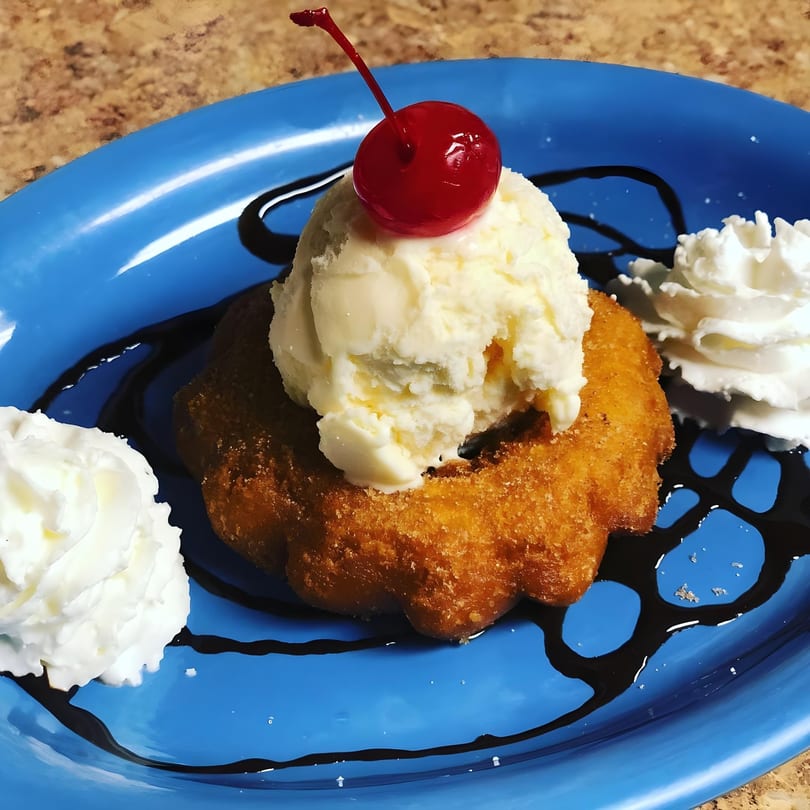 A golden-brown fried dessert topped with vanilla ice cream, a cherry, whipped cream, and chocolate syrup.