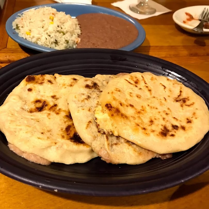 Three golden-brown pupusas on a black plate, with rice and refried beans in the background.