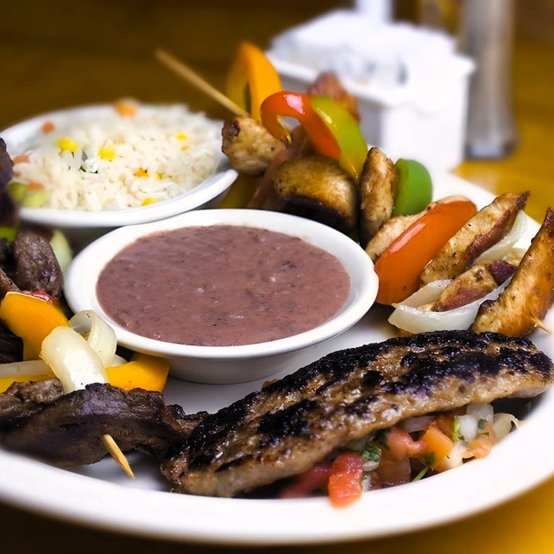 A Salvadoran dish with grilled meat, vegetable skewers, rice, and a bowl of refried beans.
